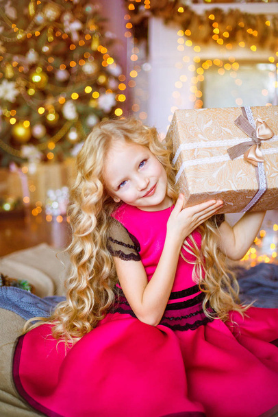 Cute little girl with long curly blond hair at home near a Christmas tree with gifts and garlands and a decorated fireplace  - Фото, изображение