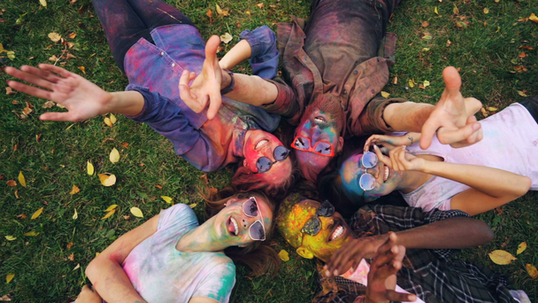 Vista superior de chicas y chicos relajados relajándose en la hierba con caras pintadas después del festival Holi mirando a la cámara, sonriendo y moviendo las manos. Recreación, diversión y concepto de naturaleza
. - Metraje, vídeo