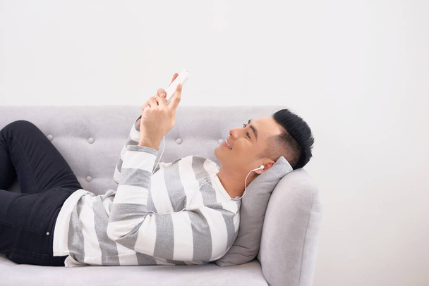 Handsome guy in earphones using a smartphone and smiling while lying on couch at home - Фото, зображення