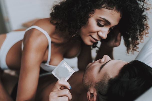 Young Beautiful Couple in Underwear Lying on Bed. Attractive Woman Sitting on Handsome Man and holding Condom. Passionate Romantic Couple about to have Sex at Home. Passion and Intimate Relationship - Foto, Bild