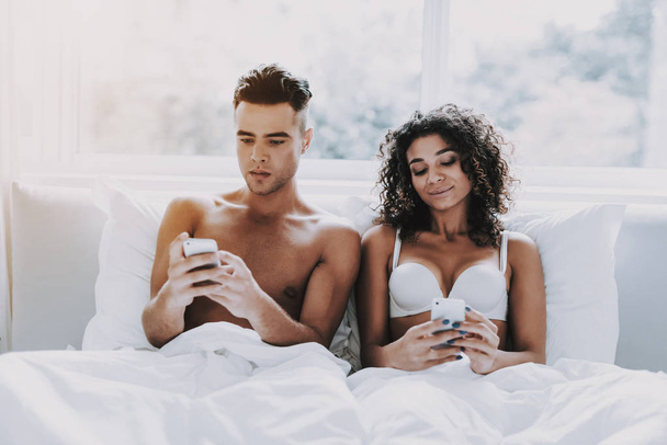 Young Beautiful Couple Using Smartphones in Bed. Attractive Woman and Handsome Man Lying in Bed Together and Using Cellphones. Happy Cute Couple in Underwear Relaxing in Bed at Home. - Foto, imagen