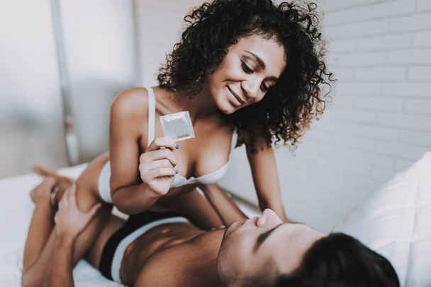 Young Beautiful Couple in Underwear Lying on Bed. Attractive Woman Sitting on Handsome Man and holding Condom. Passionate Romantic Couple about to have Sex at Home. Passion and Intimate Relationship - Фото, изображение