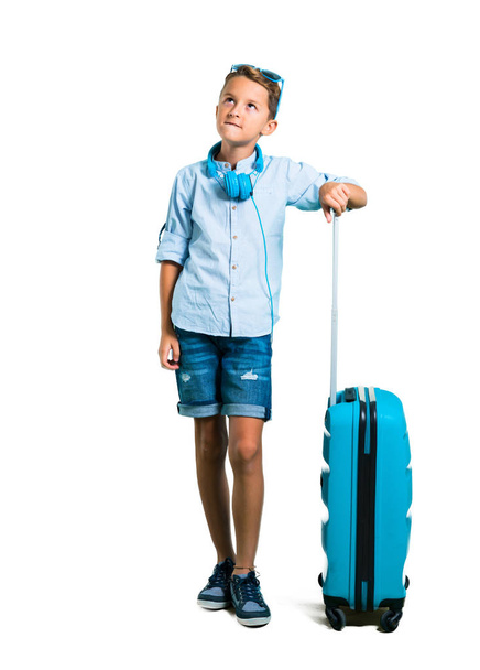 Cuerpo completo de Kid con gafas de sol y auriculares viajando con su maleta teniendo dudas y con expresión facial confusa sobre fondo blanco aislado
 - Foto, Imagen