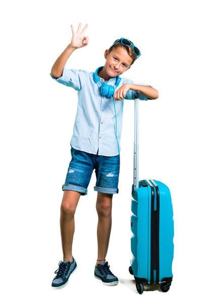 Çocuk tam vücut güneş gözlüğü ve kulaklık bavulunu bir ok işareti parmak ile izole beyaz arka plan üzerinde gösterilen ile seyahat ile - Fotoğraf, Görsel