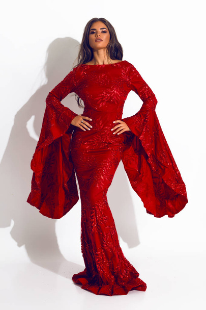Gorgeous young lady in the red long dress,  standing in the studio, hands on the waist, indoor shooting, model, beautiful face, makeup, hair style,  brunette, perfect body, event, posing, fashion look - Foto, Imagem