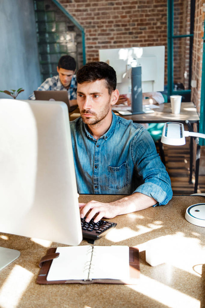 Young bearded businessman in denim shirt is sitting in office at table and is using laptop with charts, graphs and diagrams on screen. Man working.startu - Foto, afbeelding