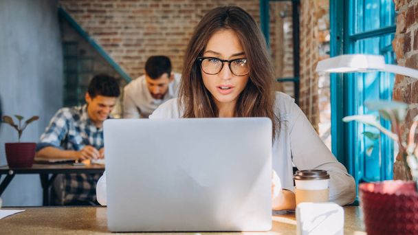 Молодая бородатая деловая женщина в джинсовой рубашке сидит в офисе за столом и использует ноутбук с графиками, графиками и диаграммами на экране. Человек работает. startu
 - Фото, изображение