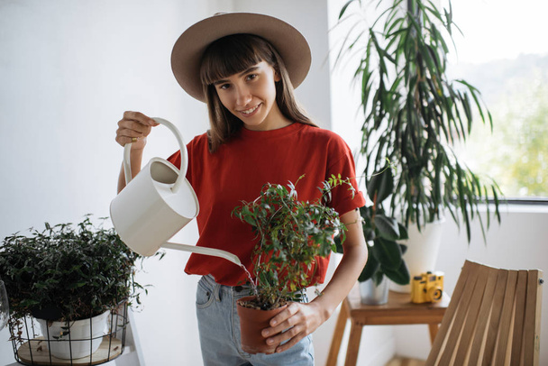 Smiling hipster gardener wearing stylish clothes and hat watering plants holding watering can in cozy green flower shop. Successful business.  - Фото, изображение