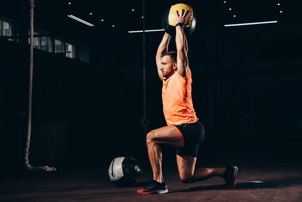 handsome fit sportsman performing lunge with medicine ball overhead in dark gym - Photo, Image
