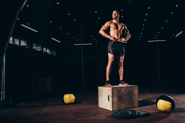 sportivo atletico senza maglietta in piedi sul cubo in palestra scura e guardando altrove
 - Foto, immagini