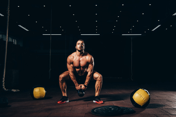 handsome athletic bodybuilder doing squats with kettlebell in dark gym - Photo, Image