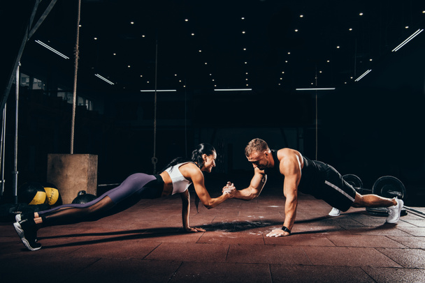 athletic sportsman and sportswoman doing push ups together and holding hands in dark gym - Photo, Image