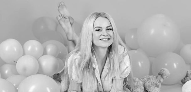Girl in pajama, domestic clothes lay near air balloons, pink background. Birthday girl concept. Blonde on smiling face relaxing with teddy bear toy. Woman cute celebrate birthday with balloons - Fotografie, Obrázek