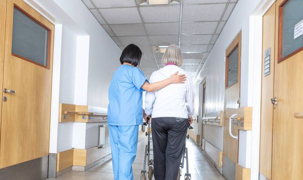 Asian doctor helping elder woman with walker in hospital hallway. - Photo, Image