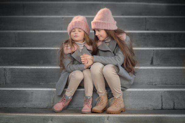 cheerful sisters wearing pink knitted hats sitting on the stairs and holding hands  - Fotografie, Obrázek