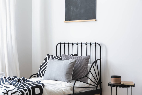 Grey pillows and patterned sheets on bed in white bedroom interior with black poster. Real photo - Fotó, kép
