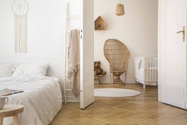 White apartment interior with herringbone parquet, double bed and open door to kid room with peacock chair, white crib and round rug on the floor - Foto, Imagem