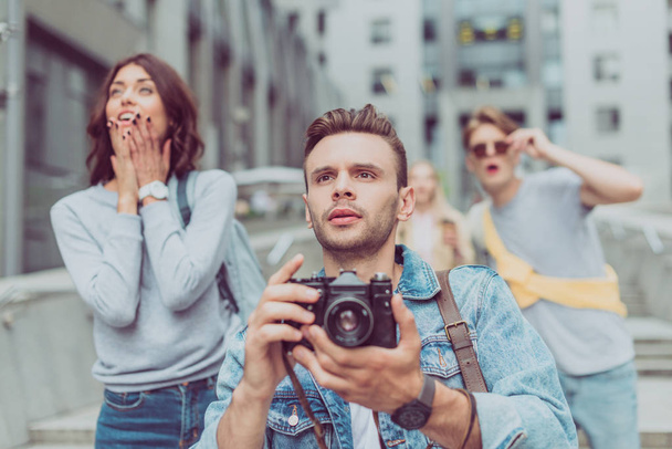 selective focus of tourist with photo camera in hands and shocked friends on street behind - 写真・画像