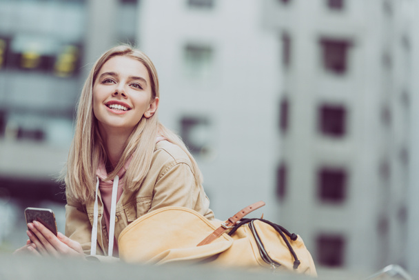 Szczęśliwa młoda kobieta blondynka z plecaka za pomocą smartfona w mieście - Zdjęcie, obraz