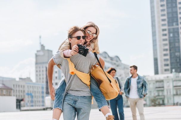 happy boyfriend giving piggyback to girlfriend with photo camera in city with friends behind - Photo, Image