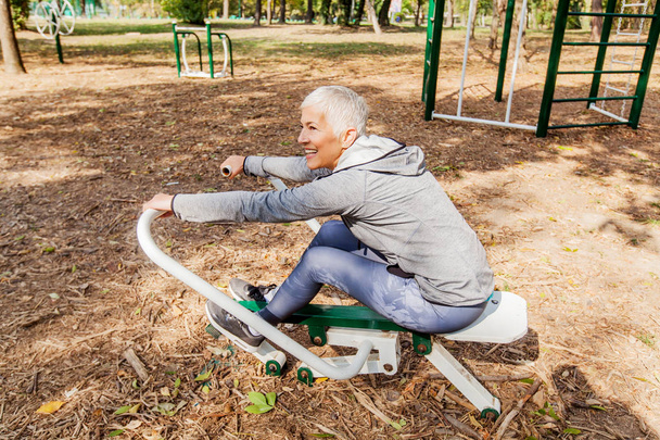 Healthy Mature Woman Practicing At Outdoor Gym, Sports Clothes, Sporty Elderly Female With Short Hair, Workout Training. - Фото, изображение