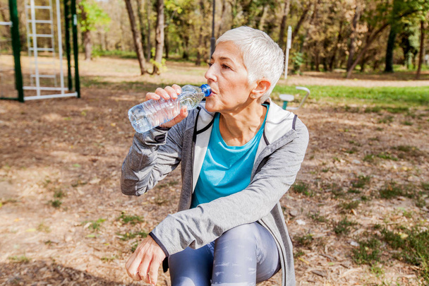 Здорова старша жінка розслабляється і п'є воду після тренування у відкритому тренажерному залі
  - Фото, зображення