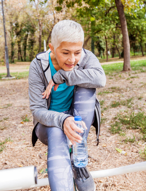 Υγιή ανώτερους γυναίκα να χαλαρώσετε και να πιείτε νερό μετά από προπόνηση στο υπαίθριο γυμναστήριο  - Φωτογραφία, εικόνα