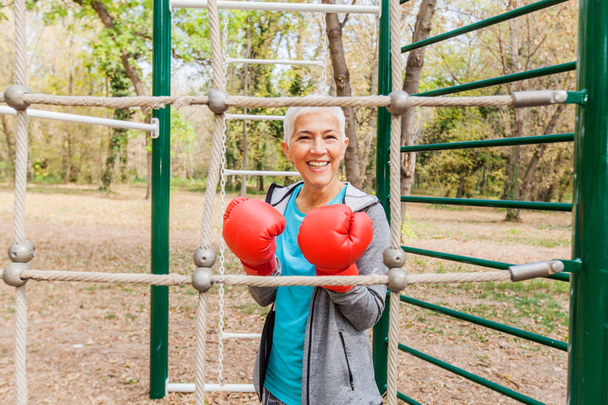 Πορτρέτο της ευτυχισμένη Fit ανώτερων γυναίκα με γάντι του μποξ στο υπαίθριο γυμναστήριο σε αθλητικά ενδύματα. Ενεργό παλιά άτομα Fitness Lifestyle - Φωτογραφία, εικόνα