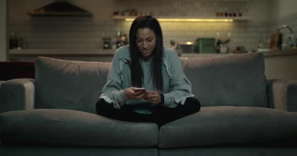 Girl sitting crossed legged on a sofa using her smartphone to chat with her friends online. - Felvétel, videó