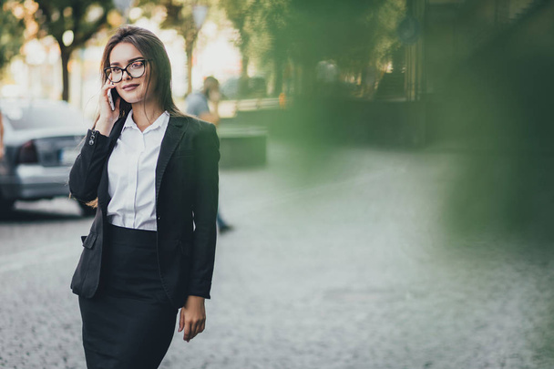 Beautiful smiling young business woman wearing formal suit, walking on city street, using smartphone, making a call - Photo, image