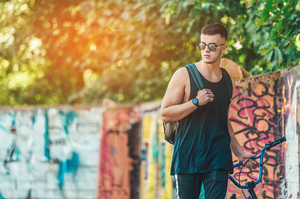 Portrait of a sporty hipster man in a black fashionable T-shirt posing with a bmx bike on a street in the summer park. Sunshine background - Fotografie, Obrázek