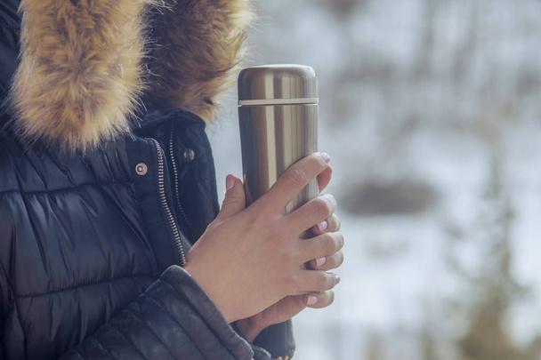 A girl in a winter jacket with a thermos of coffee or tea in her hand on the balcony in winter - Foto, imagen