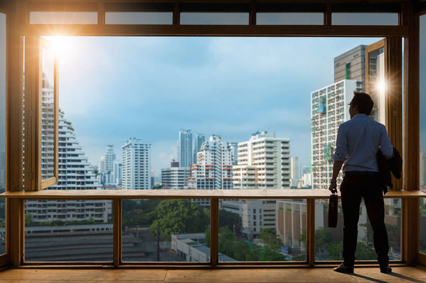 Visión conceptual, joven hombre de negocios con chaqueta cómoda traje casual de pie en la sala de trabajo cerca de la ventana de vidrio y mirando a través de los edificios modernos en el horizonte
. - Foto, Imagen