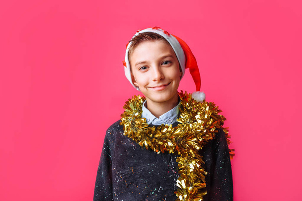 Cute teen, in Santa Claus hat and with tinsel on neck, smiling on red background - Valokuva, kuva