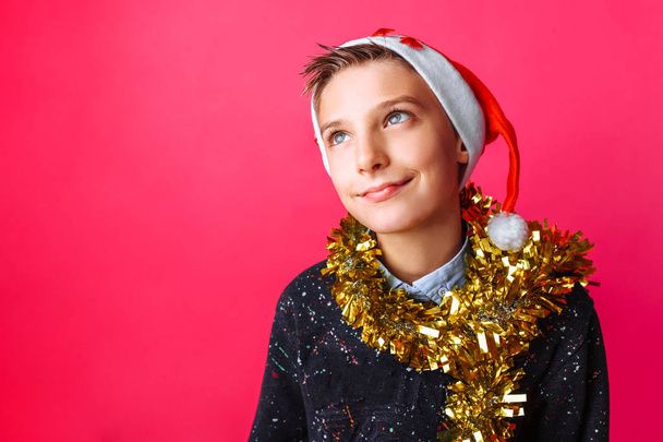 A beautiful teenager, wearing a Santa hat and with tinsel on his neck, makes plans for the upcoming New year on a red background - Fotó, kép