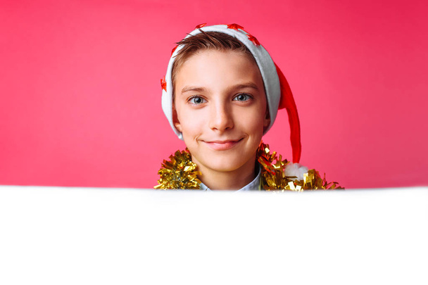 Portrait of a beautiful teen in Santa's hat and with tinsel on her neck, posing behind a white panel isolated on a red background. Teen baby holding an empty Billboard. Christmas - Foto, Bild