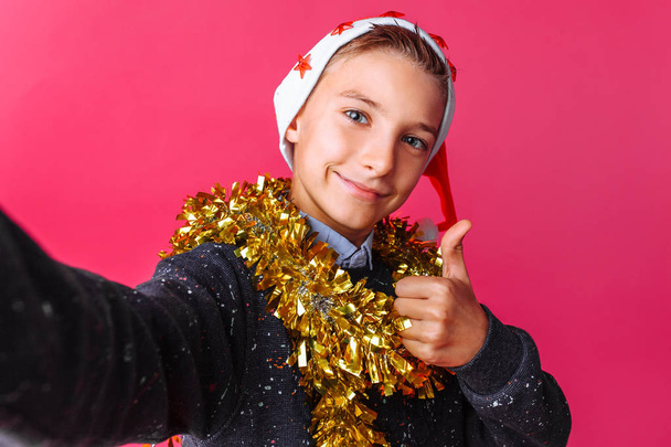 The guy a teenager, the gesture show class and makes a selfie in a Santa hat and tunnel on the neck on a red background. The concept of Christmas - Foto, Bild