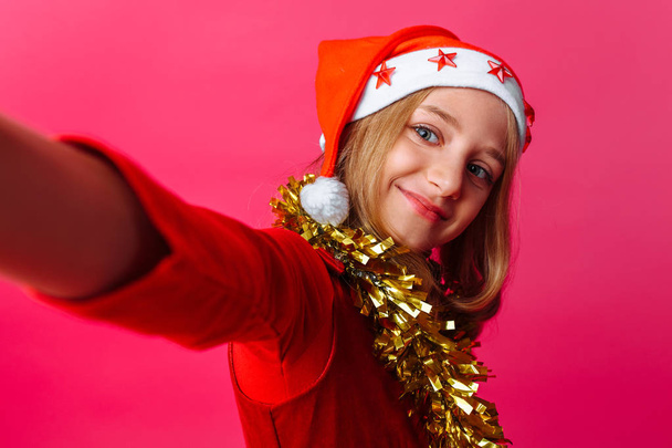 The girl makes a selfie in the hat of Santa Claus and tinsel on the neck, a schoolgirl on a red background. The Concept Of Christmas - Foto, Imagem
