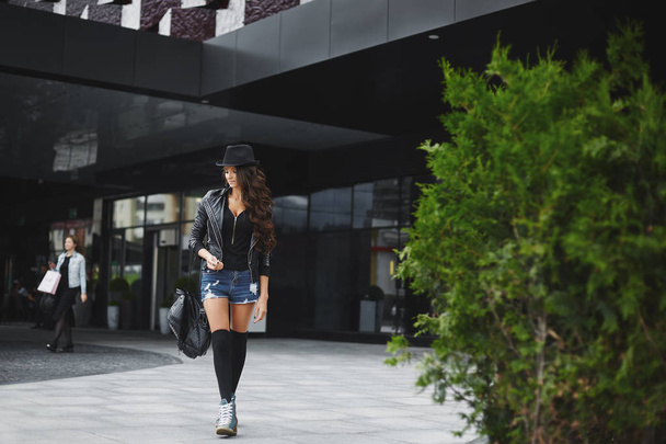Beautiful, sexy and fashionable brunette model girl with bright makeup, in the jeans shorts, in a leather jacket and in a hat, walking outdoors with a stylish black backpack in her hand - Foto, Imagem