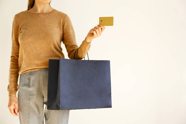 Young slim woman in classic fashionable outfit, brown sweater and loose grey pants went shopping, holding paper bags and vip golden credit / debit card. White background, copy space, close up. - Foto, Imagem