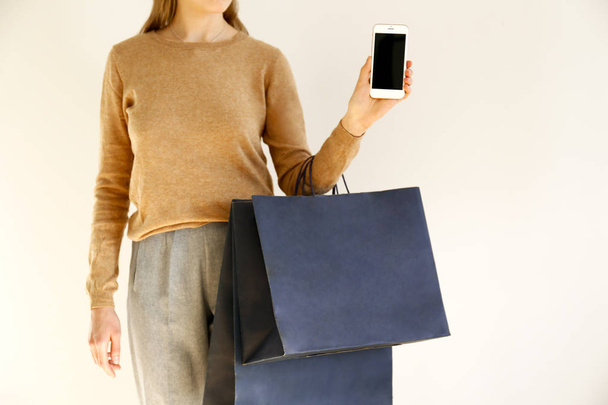 Young slim woman in classic fashionable outfit, brown sweater and loose grey pants went shopping, holding paper bags and showing blank screen smartphone. White background, copy space, close up. - Foto, afbeelding