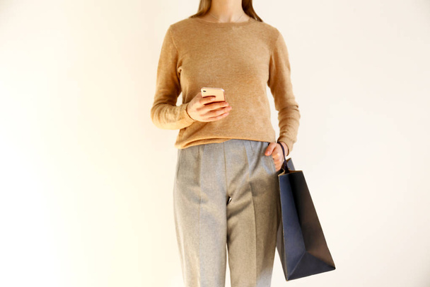 Young slim woman in classic fashionable outfit, brown sweater and loose grey pants went shopping, holding paper bags and chatting on smartphone. White background, copy space, close up. - 写真・画像