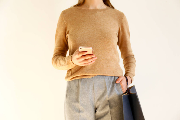 Young slim woman in classic fashionable outfit, brown sweater and loose grey pants went shopping, holding paper bags and chatting on smartphone. White background, copy space, close up. - Φωτογραφία, εικόνα