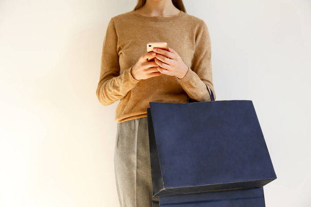 Young slim woman in classic fashionable outfit, brown sweater and loose grey pants went shopping, holding paper bags and chatting on smartphone. White background, copy space, close up. - Zdjęcie, obraz