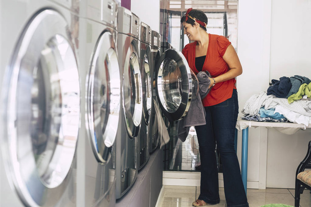 brunette woman washing and cleaning some dresses and clothes at laundry mat service automatic machine - Photo, image