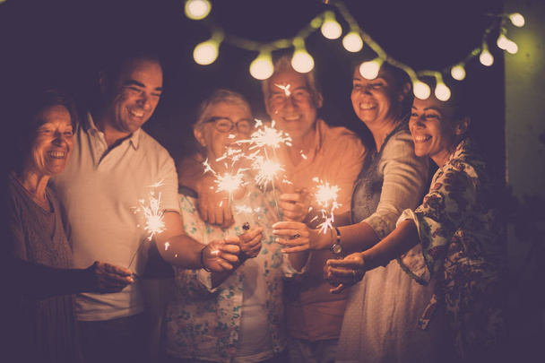 skupina bělochů přátelé s různým věkem slaví společně narozeniny nebo Silvestra v noci venku doma. světla a jiskry s veselými ženami a muži baví v přátelství - Fotografie, Obrázek