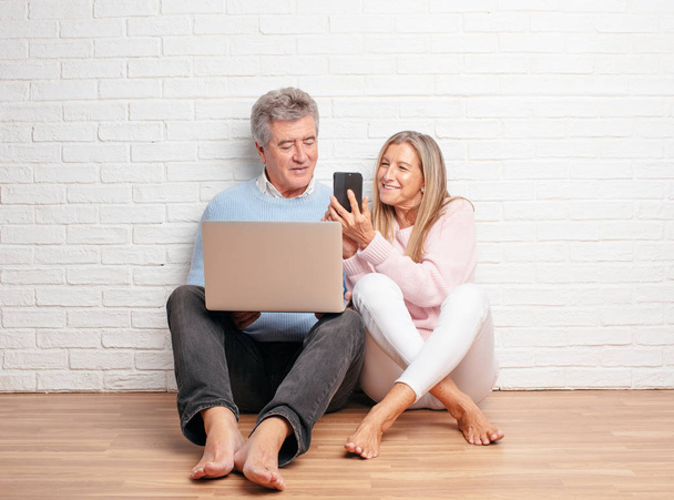pretty senior couple sitting on their house floor with a laptop - Фото, изображение