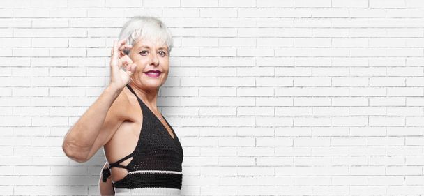 senior cool woman making an "alright" or "okay" gesture approvingly with hand, looking happy and satisfied, with a broad smile. Positive check sign. Lateral or side view. - Фото, изображение