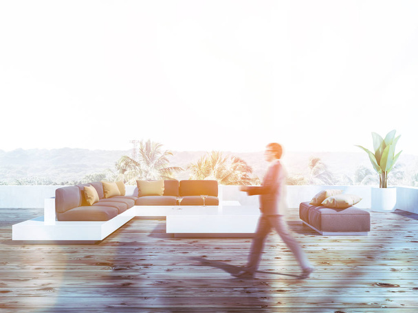 Man walking on his balcony with wooden floor, large white sofa with cushions and coffee table. Toned image blurred - 写真・画像