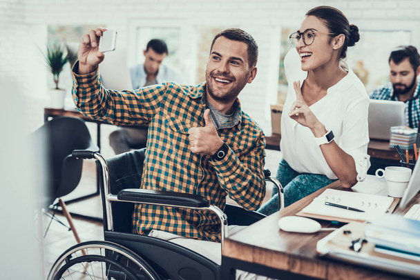 Man on Wheelchair Taking Selfie with Young Girl. Using Smartphone. Teamwork in Office. Young Worker. Smiling Woman. Woman in Glasses. Using Digital Device. Disabled Young Man. Man on Wheelchair. - 写真・画像